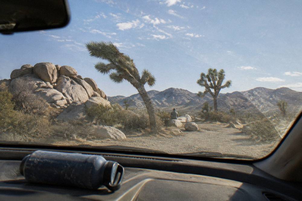 A sedan parked at a desert pullout beside tall Joshua trees and stacked boulders