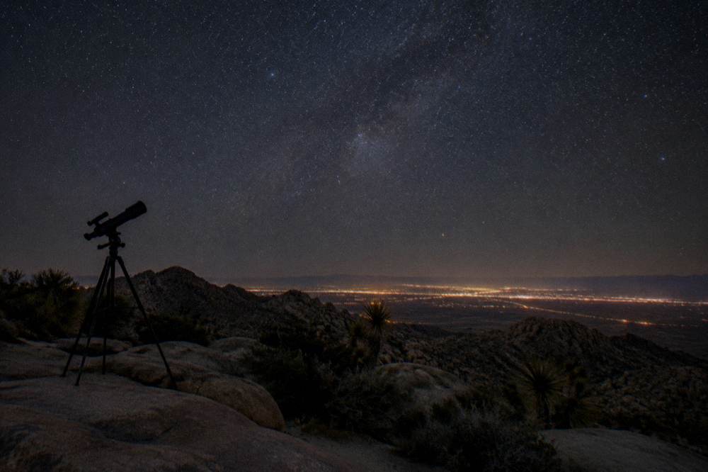 Telescope silhouetted against a star-filled night sky over dark desert ground
