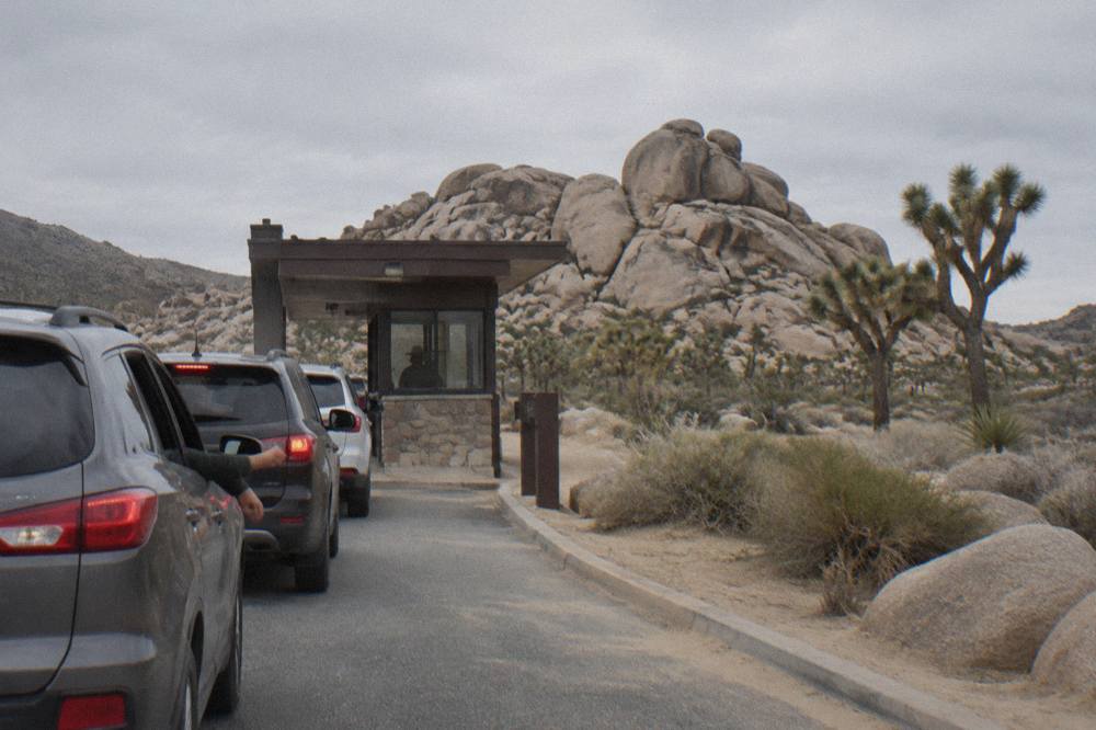 A line of cars waiting at a small desert entrance station building beneath tall rocks