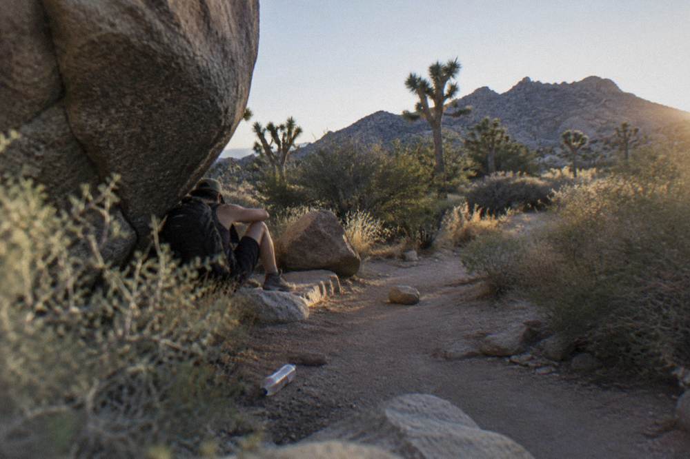 A lone hiker resting in the shade of a massive boulder beside a dusty trail