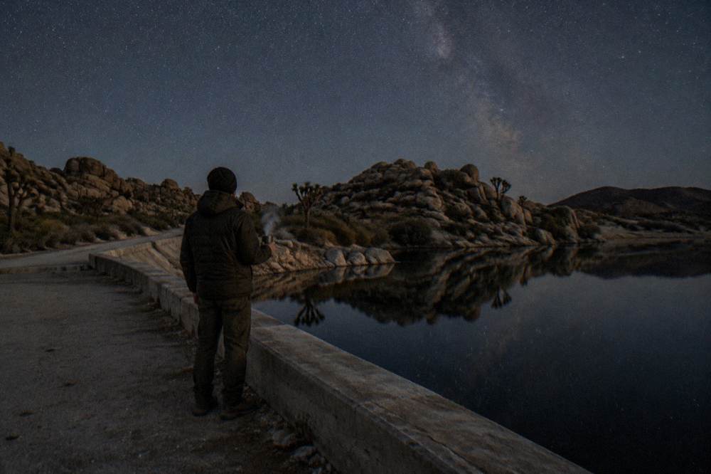 A person silhouette under a dark desert sky filled with stars and a faint Milky Way band