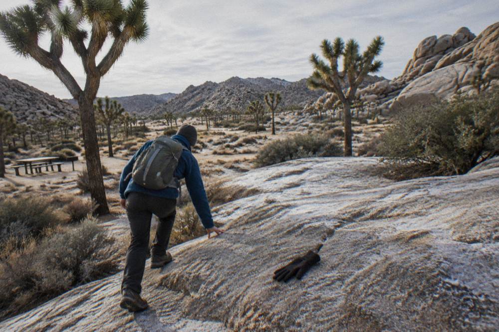 A hiker in layers walking a frosty granite trail among Joshua trees in morning light