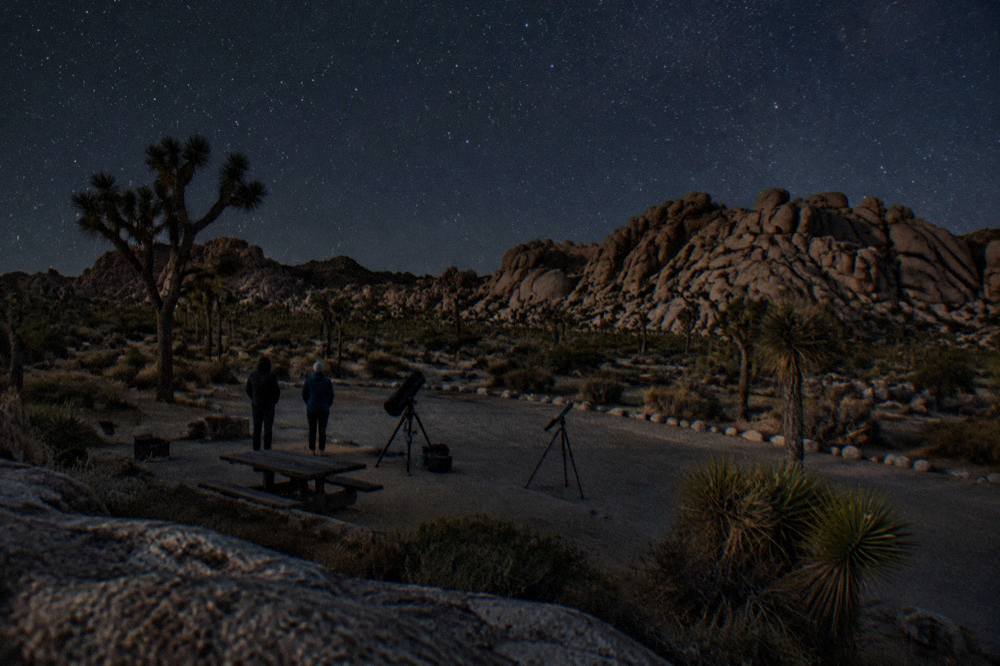 A dark desert parking area with a group of telescopes set up under a starry sky