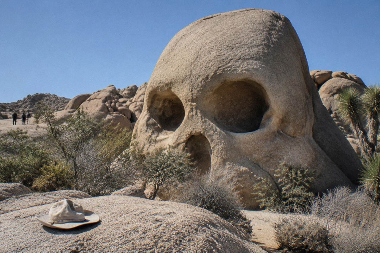 A large rounded boulder formation with two hollowed eye-like openings surrounded by desert rocks and scrub