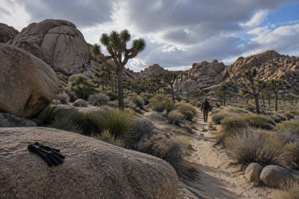 A hiker walking a sandy trail between granite rock formations and joshua trees
